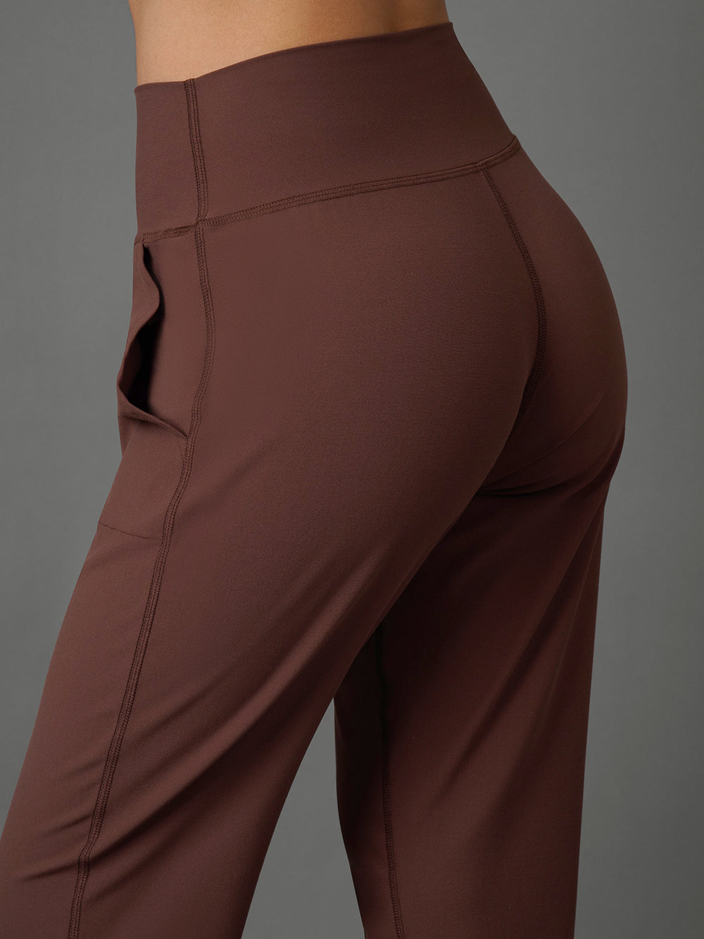 Close-up of a model wearing high-waisted brown activewear leggings with side pockets, shown from a rear three-quarter angle against a solid gray background.