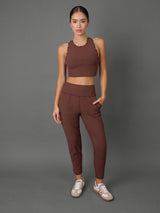 A woman modeling a matching brown activewear set consisting of a halter crop top with scalloped edges and high-waisted jogger-style pants, paired with white sneakers, against a solid gray studio background.