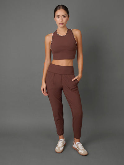 A woman modeling a matching brown activewear set consisting of a halter crop top with scalloped edges and high-waisted jogger-style pants, paired with white sneakers, against a solid gray studio background.